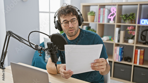 Handsome hispanic man with beard recording in a radio studio, wearing headphones and reading a script.