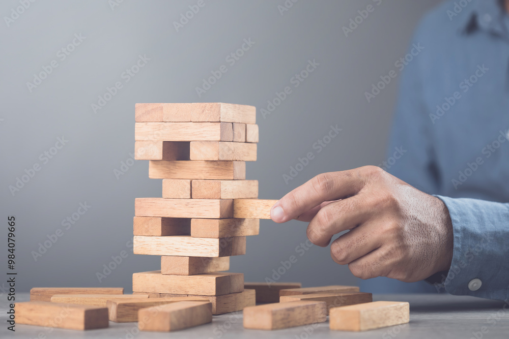 Businessman contemplates his next move in a stacking tower block game on a table against a gray wall background. Symbolizes strategic thinking, planning, and decision-making in business and life Businessman contemplates his next move in a stacking tower block game on a table against a gray wall background. Symbolizes strategic thinking, planning, and decision-making in business and life