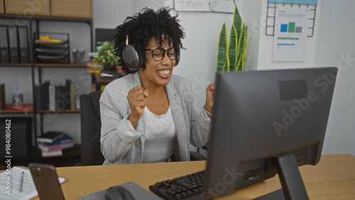 Young african american woman celebrating success in modern office environment with headphones on at her workplace desk.