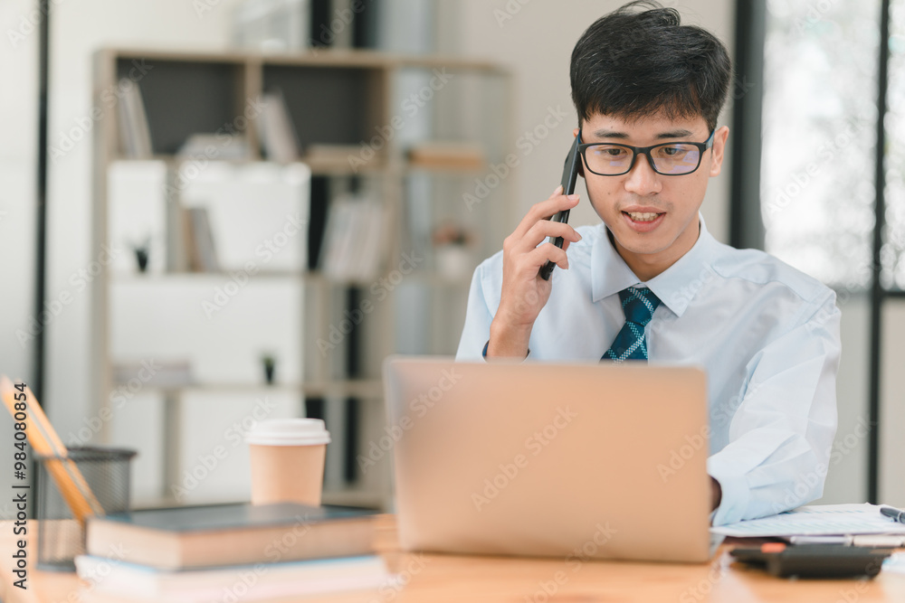 A young professional works at a desk, talking on the phone while using a laptop, surrounded by books and a coffee cup, in a modern office setting. A young professional works at a desk, talking on the phone while using a laptop, surrounded by books and a coffee cup, in a modern office setting.
