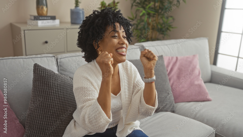 A beautiful young african american woman with curly hair sits on a comfortable couch in her living room, smiling and celebrating with excitement. A beautiful young african american woman with curly hair sits on a comfortable couch in her living room, smiling and celebrating with excitement.