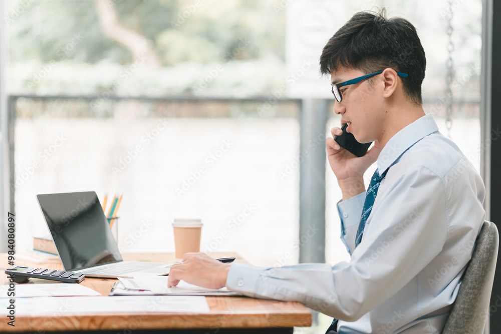 A professional young man is sitting at a desk, talking on the phone while working on a laptop, with a coffee cup beside him in a bright office setting. A professional young man is sitting at a desk, talking on the phone while working on a laptop, with a coffee cup beside him in a bright office setting.