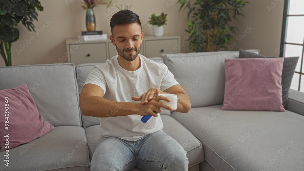 Handsome hispanic man applying lotion in a comfortable living room, showcasing a cozy home interior with soft furnishings and plants around. Handsome hispanic man applying lotion in a comfortable living room, showcasing a cozy home interior with soft furnishings and plants around.