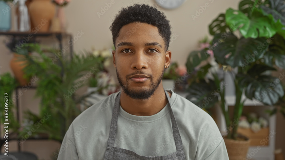 Handsome african man wearing an apron standing in a green indoor flower shop environment Handsome african man wearing an apron standing in a green indoor flower shop environment