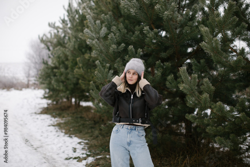 A woman in a short brown sheepskin coat and jeans stands near evergreen trees in winter. photo