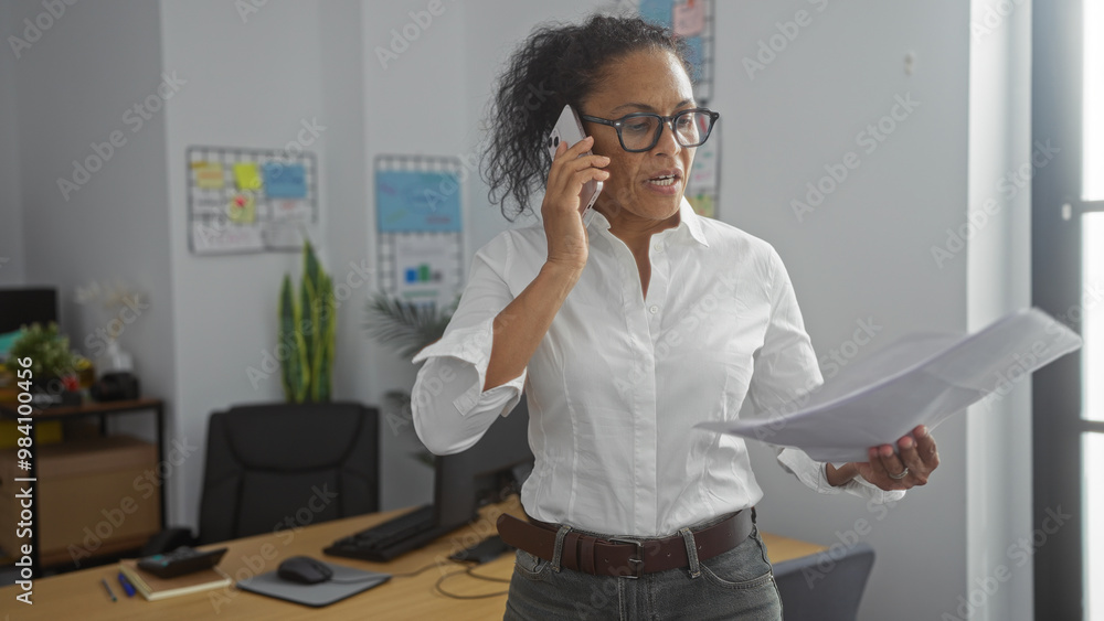Mature woman talking on the phone and holding papers in an office setting, surrounded by work supplies and decor Mature woman talking on the phone and holding papers in an office setting, surrounded by work supplies and decor