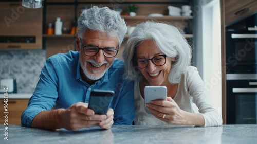 Happy senior couple looking at a smartphone, lying on a kitchen counter and smiling while watching a video or reading a message from family online in a modern home interior. The old man with grey hair