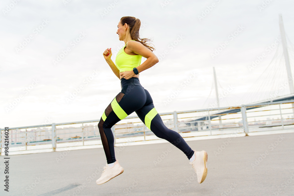 Fototapeta premium A Woman in Bright Workout Gear Runs Along a Waterfront Pathway During Early Morning Hours, Enjoying Her Fitness Routine