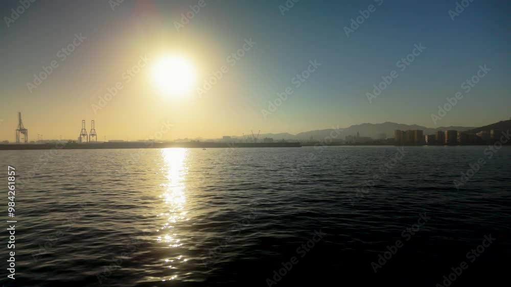 A serene sunset over the water at Malaga port, with the sun casting a golden reflection on the sea and cranes visible in the distance A serene sunset over the water at Malaga port, with the sun casting a golden reflection on the sea and cranes visible in the distance