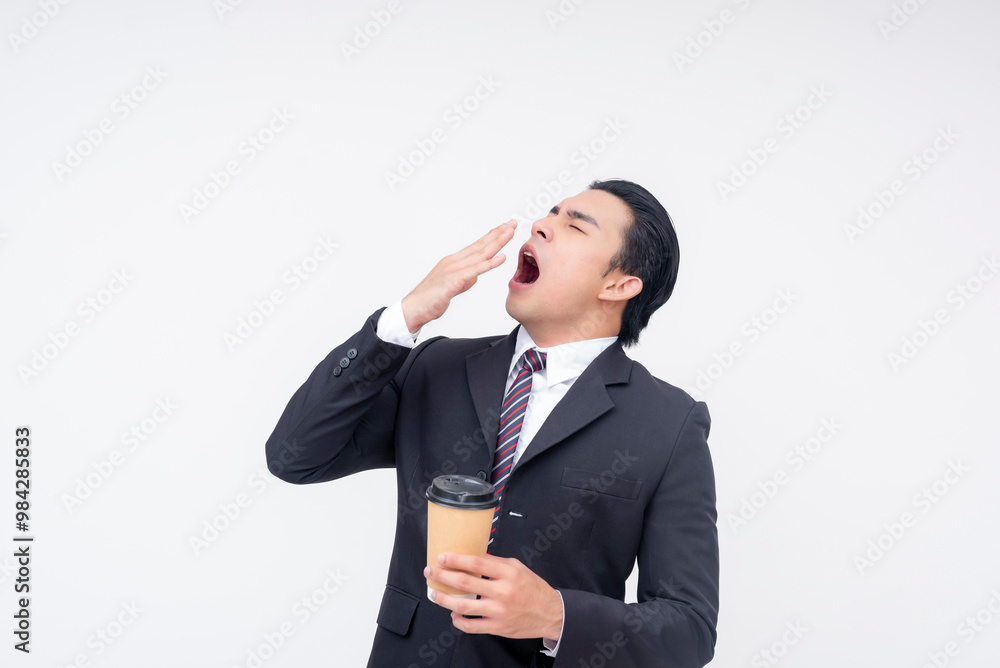 A tired young asian businessman in a formal suit yawning while holding a cup of coffee. Isolated on a white background. A tired young asian businessman in a formal suit yawning while holding a cup of coffee. Isolated on a white background.
