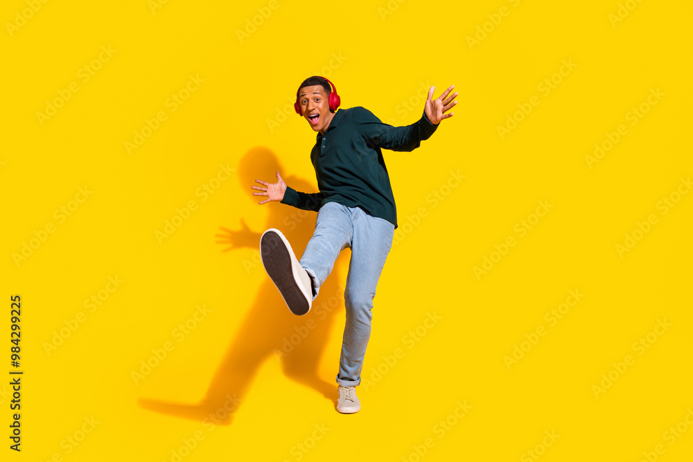 Full length photo of attractive young man headphones dance have fun dressed stylish green clothes isolated on yellow color background Full length photo of attractive young man headphones dance have fun dressed stylish green clothes isolated on yellow color background