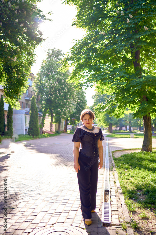 Fototapeta premium a medium size woman in a black corset and wide leg trousers walks through the city in the morning light