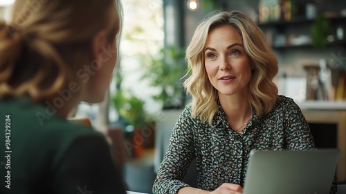 Two happy busy business women of young and middle age talking in green cozy office sitting at desk Professional ladies executives having conversation using laptop at work Authentic can : Generative AI