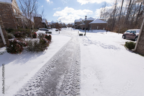 A quiet suburban neighborhood blanketed in snow, showing tire tracks on a driveway under a bright, sunny sky