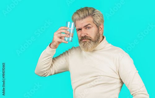 Man take some pills, holds glass of water, isolated on blue. Berded man taking drugs to releave headache. Man taking pill against headache. Male taking a pill with a glass of water photo