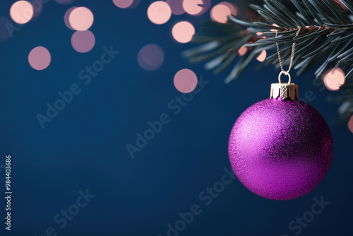 Purple Christmas ornament hanging from a pine branch with warm bokeh lights in the background, set against a deep blue festive backdrop.