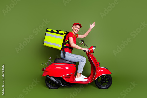 Full body photo of mature woman delivery employee thermo bag ride scooter wave hand wear red uniform workwear isolated on khaki background