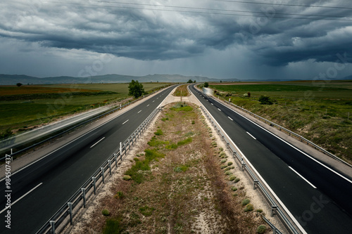 Pamnoramic view of a divided highway cutting through a rural landscape with fields and distant hills under a cloudy sky.