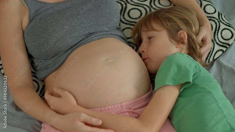 A pregnant woman and her elder son lie on a bed, sharing a tender moment. The boy hugs his mother's belly and talks to his unborn sibling, capturing the beauty of family bonds and the anticipation of A pregnant woman and her elder son lie on a bed, sharing a tender moment. The boy hugs his mother's belly and talks to his unborn sibling, capturing the beauty of family bonds and the anticipation of