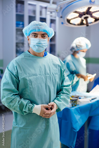 A man in a blue surgical gown stands in front of a surgical table. He is wearing a blue hat and a blue mask. Another person is standing behind him