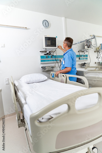 A man in a blue shirt is standing in front of a hospital bed. He is checking the patient's vitals