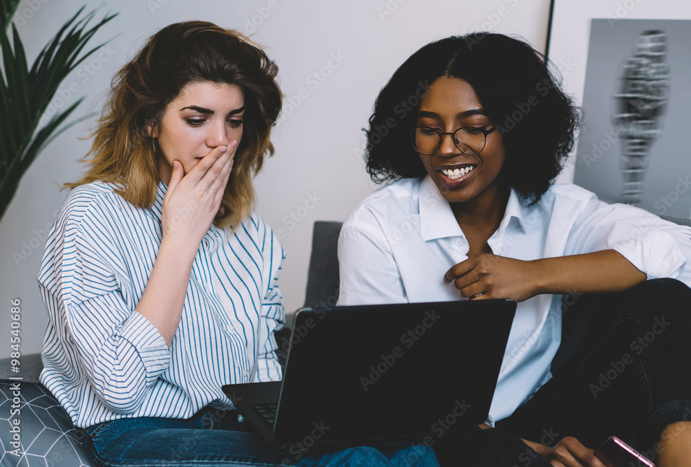 Smiling african american young woman and caucasian hipster girl shocked with last fashion news on website, millennial diverse friends watching video on laptop computer using high home internet Smiling african american young woman and caucasian hipster girl shocked with last fashion news on website, millennial diverse friends watching video on laptop computer using high home internet
