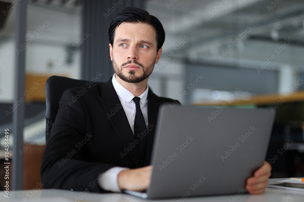 A man in a suit is sitting at a desk with a laptop in front of him. He is focused on his work, possibly preparing for a meeting or working on a project. Concept of professionalism and concentration A man in a suit is sitting at a desk with a laptop in front of him. He is focused on his work, possibly preparing for a meeting or working on a project. Concept of professionalism and concentration