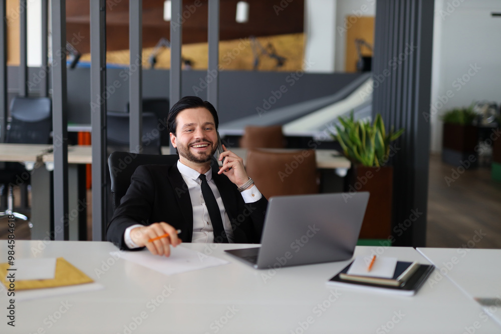 A man in a suit is talking on his cell phone while sitting at a desk. He is smiling and he is in a good mood A man in a suit is talking on his cell phone while sitting at a desk. He is smiling and he is in a good mood
