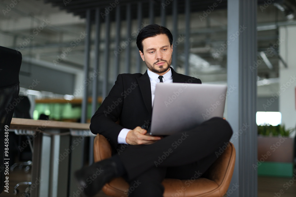 A man in a suit is sitting in a chair with a laptop in front of him. He is focused on his work, possibly typing or browsing the internet. Concept of professionalism and productivity A man in a suit is sitting in a chair with a laptop in front of him. He is focused on his work, possibly typing or browsing the internet. Concept of professionalism and productivity