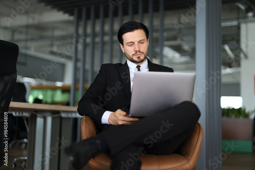 A man in a suit is sitting in a chair with a laptop in front of him. He is focused on his work, possibly typing or browsing the internet. Concept of professionalism and productivity