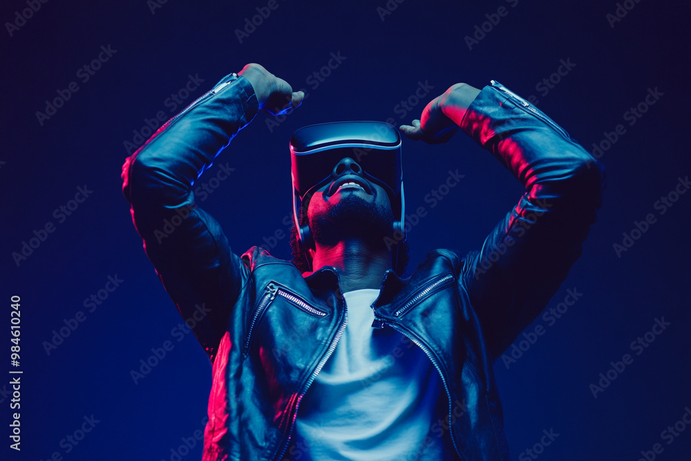 A man wearing a leather jacket and a white T-shirt is standing with his hands raised above his head. A man wearing a leather jacket and a white T-shirt is standing with his hands raised above his head.