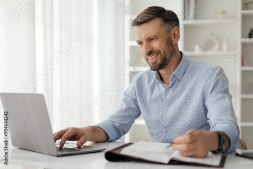 The man focuses intently on his laptop, taking notes in a notebook. Sunlight floods the modern space, highlighting his productive atmosphere and diligent effort.