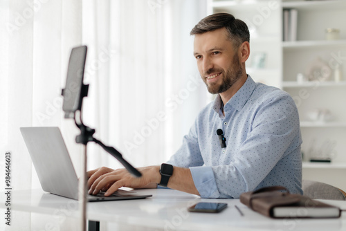 A man is seated at a bright desk, focused on his laptop as he prepares for a video call. He appears engaged and ready to communicate professionally.