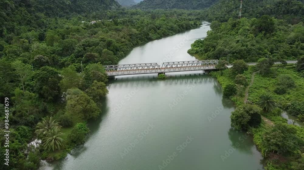 The video showcases a drone flying above a river with turquoise-colored water. As it glides along the river's course, the drone passes over an iron frame bridge The video showcases a drone flying above a river with turquoise-colored water. As it glides along the river's course, the drone passes over an iron frame bridge