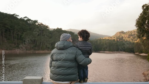 A tender moment as a father holds his child, both overlooking a calm lake surrounded by forests at dusk.