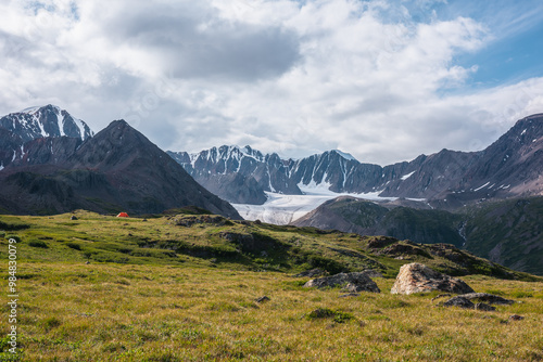 Dramatic scenery with orange tent in wide alpine valley among green hills and rocks with view to big glacier tongue and large snow-capped mountain range under clouds in blue sky in changeable weather.