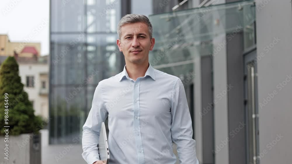 A confident mature businessman walks towards the camera outside the office. He is smiling slightly and holding a laptop. He is wearing a button down shirt and dress pants. A confident mature businessman walks towards the camera outside the office. He is smiling slightly and holding a laptop. He is wearing a button down shirt and dress pants.