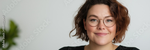 Close-up portrait of a cheerful, attractive woman with short, curly brown hair and stylish round eyeglasses, smiling genuinely at the camera in a modern office setting