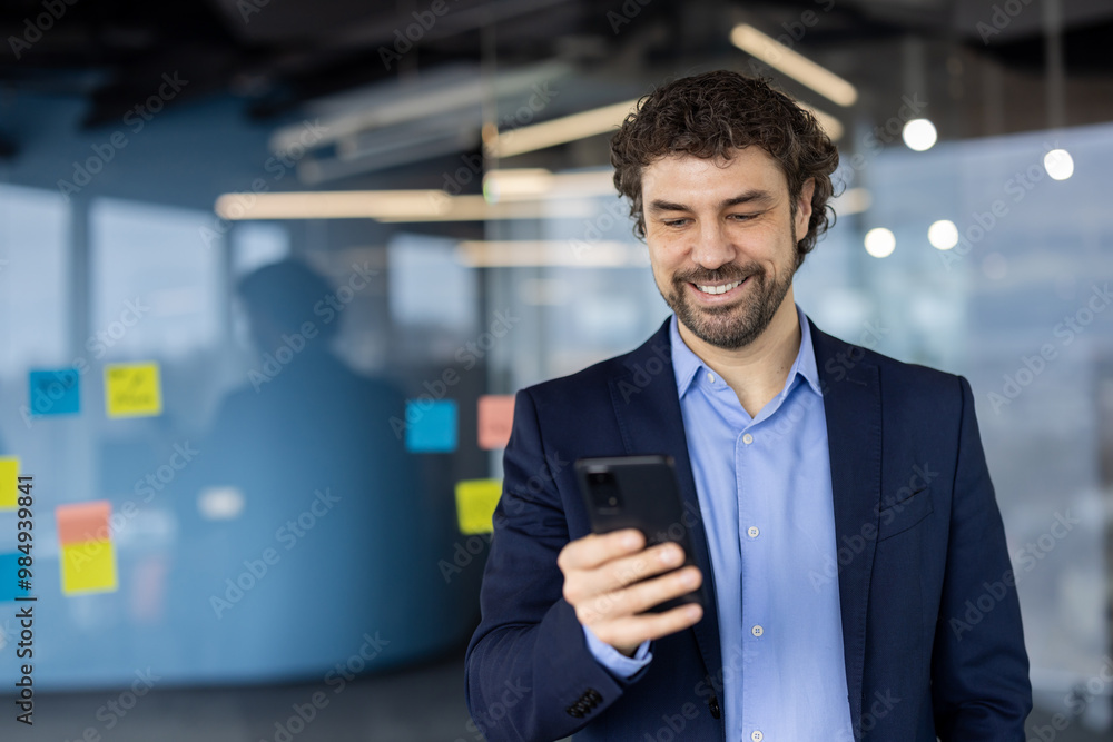 Businessman wearing suit using smartphone in modern office. Man smiling, engaging with phone for work communication or tasks. Office environment with note-covered glass board in background. Businessman wearing suit using smartphone in modern office. Man smiling, engaging with phone for work communication or tasks. Office environment with note-covered glass board in background.
