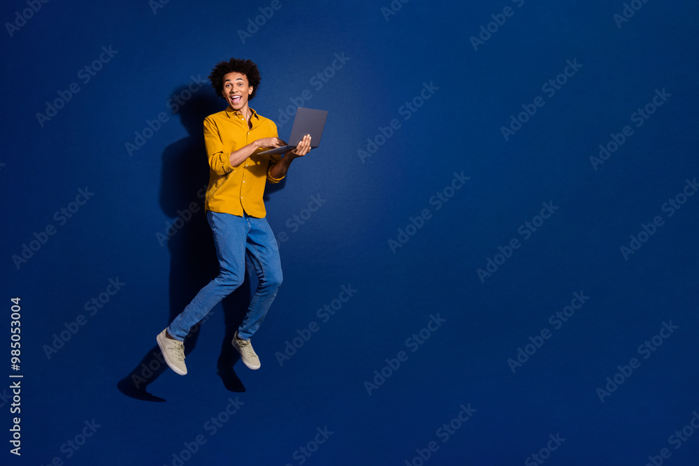 Full body portrait of nice young man jump laptop empty space wear yellow shirt isolated on dark blue color background Full body portrait of nice young man jump laptop empty space wear yellow shirt isolated on dark blue color background