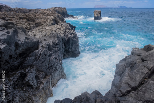 a cliff of black volcanic rock with a sea of turquoise water with waves lapping against the rocks, a picturesque rock rising out of the water