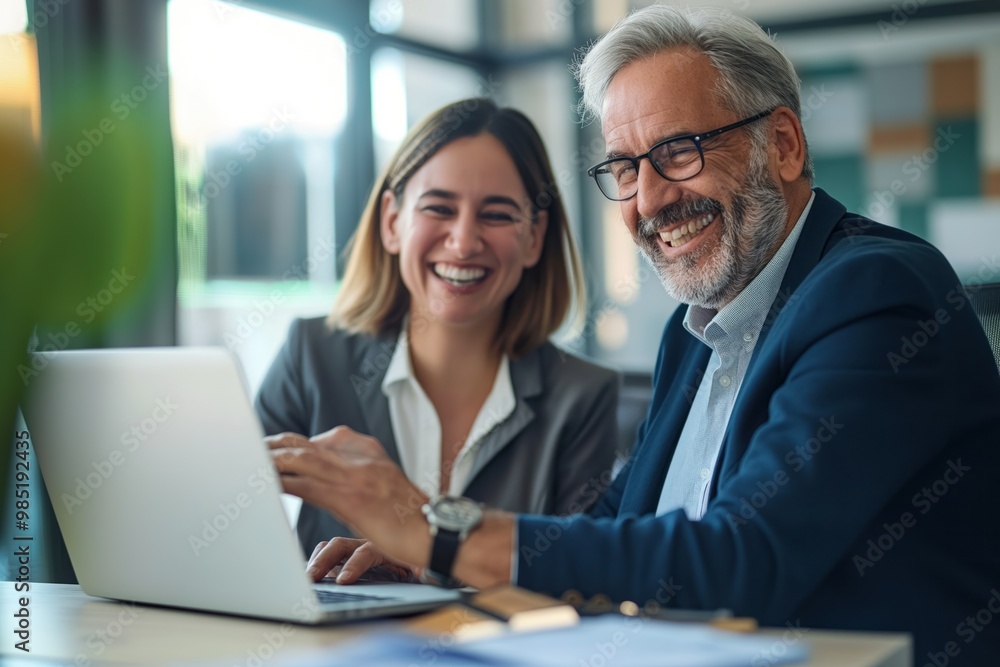 Obraz premium Two middle-aged business professionals smiling confidently at office desk. Man and woman work together, use laptop for project. Happy and successful colleagues collaborate, manage business.