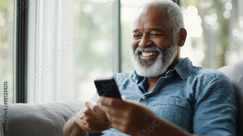 A smiling man with a beard sits comfortably on a couch, enjoying and looking at his smartphone near a bright window.