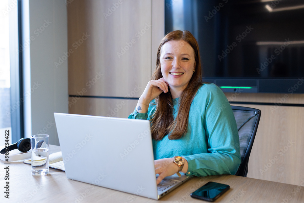Smiling woman working on laptop in office, phone and headphones on desk Smiling woman working on laptop in office, phone and headphones on desk