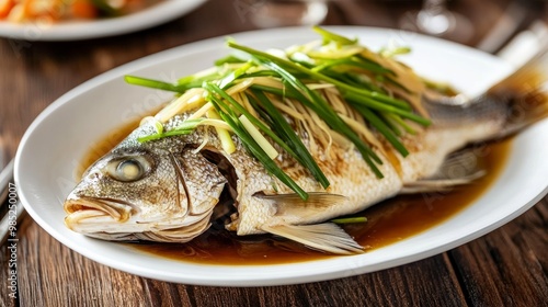 A whole steamed fish garnished with ginger, green onions, and soy sauce, displayed on a white porcelain dish at a festive table.