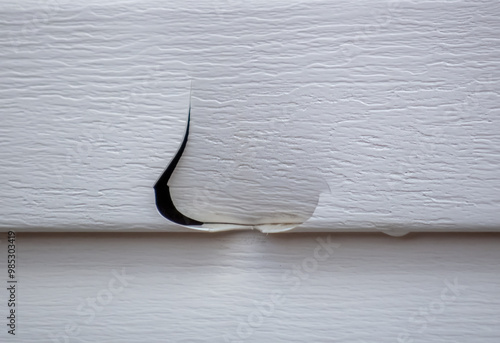 A small, tear-shaped hole has appeared in a white vinyl siding panel, indicating potential hail damage or structural issues.
