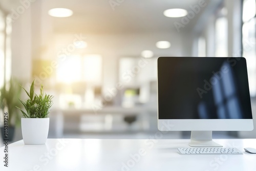 Close up of a white computer desktop with a monitor, keyboard and mouse with a potted plant in the foreground and a blurred office in the background.