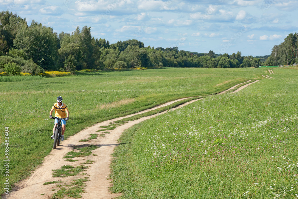 Cyclist in helmet with equipment on electric bicycle rides along path in field in countryside. Sports and health. Summer hobby. Cyclist in helmet with equipment on electric bicycle rides along path in field in countryside. Sports and health. Summer hobby.