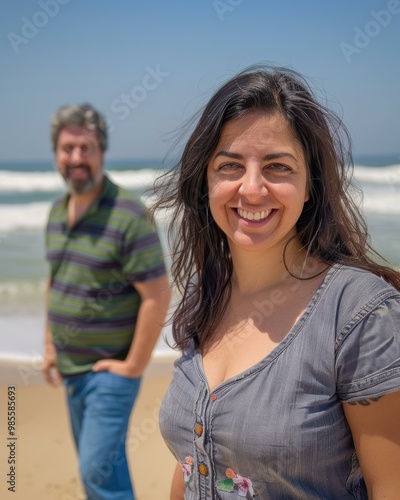 A woman smiles at the camera while standing on the beach with a man behind her. AI.