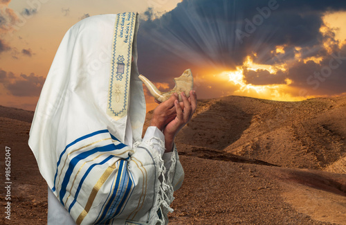Man in traditional Jewish prayer attire including a tallit with blue stripes blowing a shofar at sunset in a desert landscape. Rays of sunlight pierce through the clouds, creating a spiritual ambiance